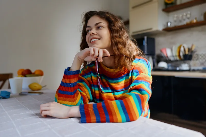 Cheerful woman indoors at home, wearing a colorful striped sweater, relaxed pose at a dining table, soft natural light, cozy kitchen backdrop, casual lifestyle moment