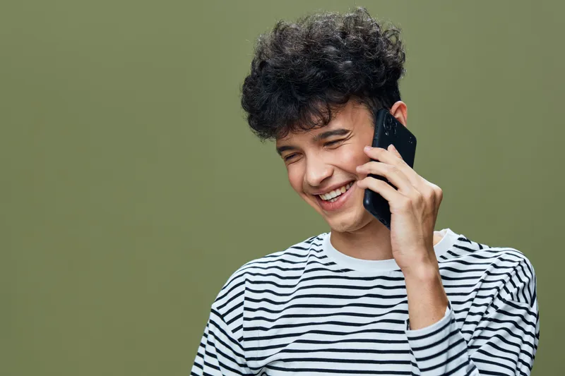 Smiling young man talking phone, casual striped shirt, curly hair, cheerful expression, green background, modern lifestyle communication.