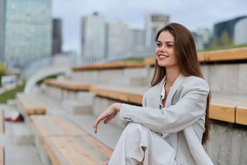 Businesswoman smiling outdoors, dressed in a light gray suit, represents confidence and professionalism in an urban setting, ideal for corporate themes.
