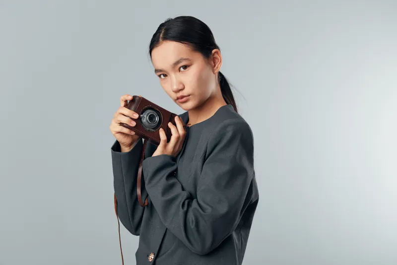 Young woman holding vintage camera, portrait in studio with gray background. Female photographer looking at viewer, casual outfit and focused expression with retro style camera.