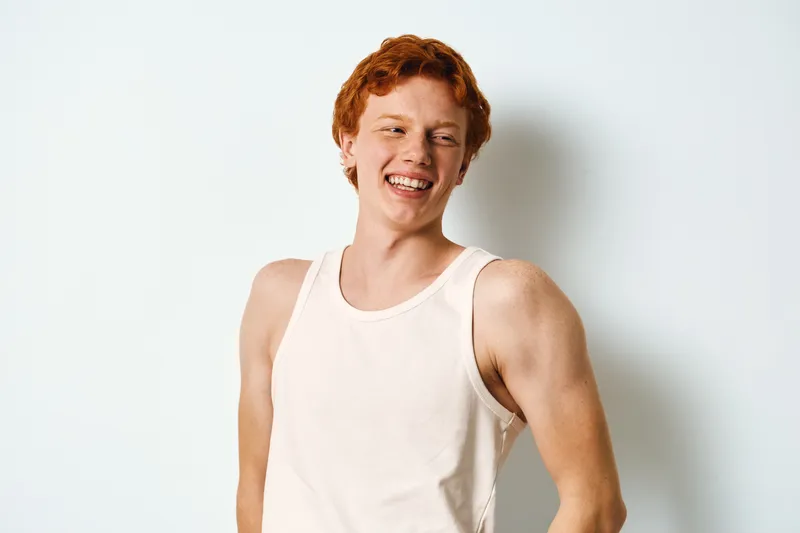 Young man with red hair wearing white sleeveless shirt smiling and laughing against white background in casual style. Male model looking happy and relaxed in studio photography.