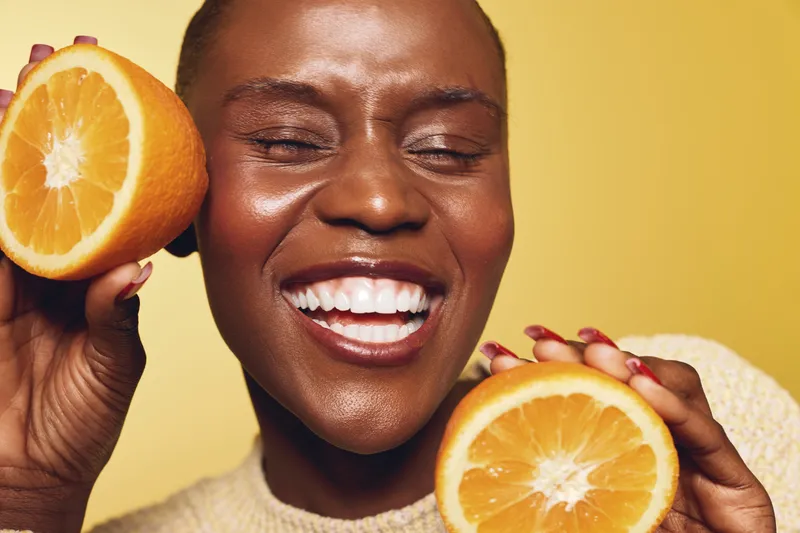Smiling dark skinned African woman holds two orange halves close to her face, eyes closed and teeth visible. Concept of healthy lifestyle, joy, natural beauty, and fresh citrus fruit.