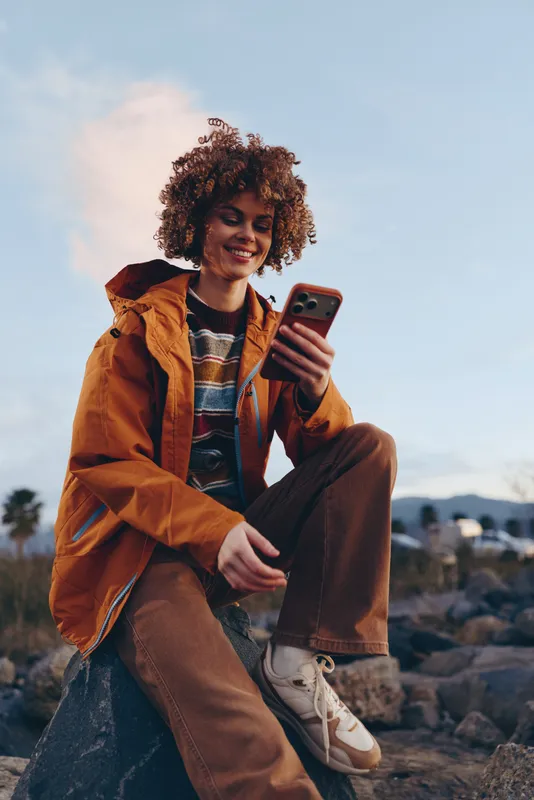 Woman lifestyle outdoors smiling with curly hair holding smartphone in orange jacket, casual travel mood, golden evening sky, soft light, relaxed pose on rocks near shore, palm trees