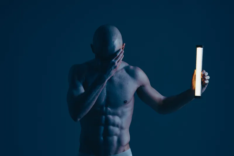 A contemplative man holding a light source, showcasing his muscular physique in a dark setting. The emphasis is on shadows and light contrasts, creating a captivating atmosphere.