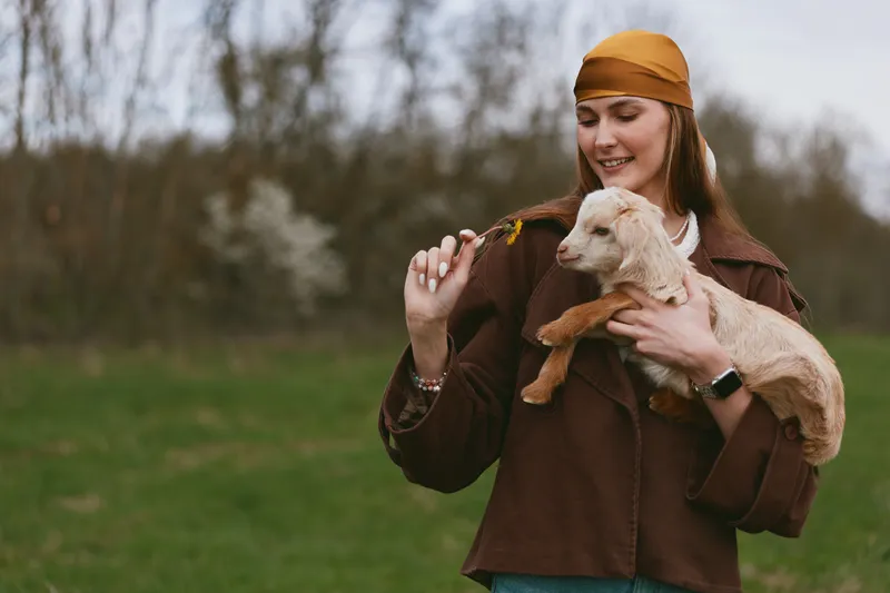 A smiling woman outdoors holds a young goat kid, gently offering a flower in a sunny, natural setting. A candid lifestyle moment centered on companionship with animals in a park-like field with soft