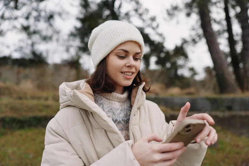 Woman in a warm coat and beanie outdoors, using a smartphone, relaxed and focused, capturing a candid moment in a chilly park setting during late autumn