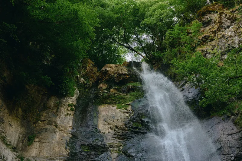 Beautiful natural waterfall cascading down rocky cliffs surrounded by lush green trees, creating a serene and refreshing outdoor landscape in a forest setting.