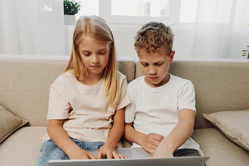 Two focused children sitting on a beige sofa using a laptop together in a bright room with natural light. Concept of learning, technology, teamwork, and childhood development.