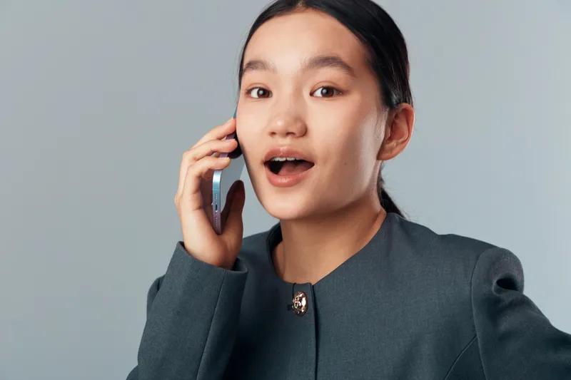 Young woman talking on smartphone with surprised expression in studio. Portrait of female person wearing dark jacket managing phone call with open mouth and attentive eyes on grey background.