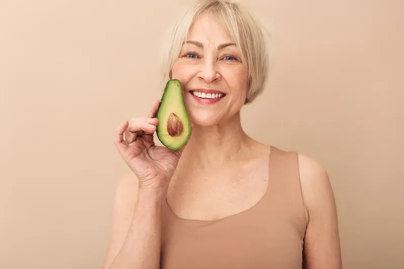 Smiling mature woman with short blonde hair holds half avocado near face, wearing sleeveless top. Beige background emphasizes natural skincare, healthy lifestyle, and wellness concept.