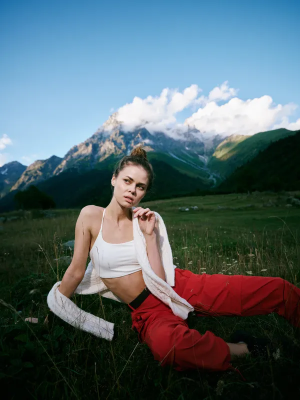 Outdoor fashion, mountain landscape, woman, casual wear, red pants, white top, cardigan, nature, alpine scenery, grassy meadow, relaxed pose, styling, blue sky, valley greenery, lifestyle serenity,