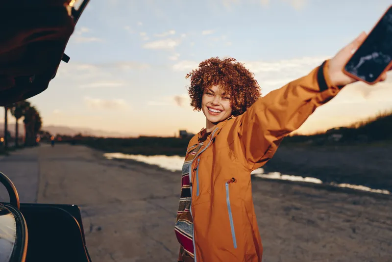 Woman lifestyle outdoors, smiling curly hair, orange jacket, arms open in soft breeze, golden sunset light, relaxed moment near street and open sky, candid travel style