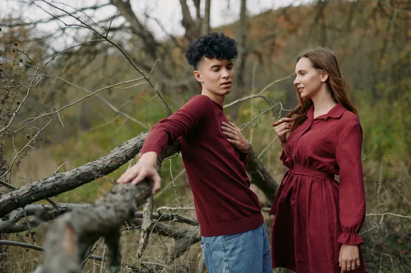A candid outdoor portrait of a young couple in matching burgundy outfits leaning on fallen branches, conveying natural interaction, genuine emotion, and lived authenticity in a simple moment.