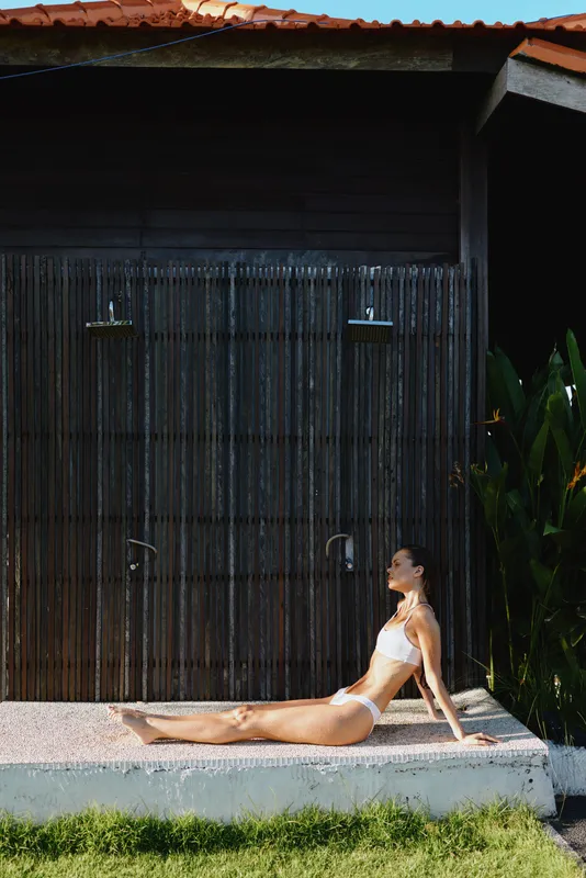A woman in a white bikini sits on a concrete ledge beside a dark wooden door, sunlit garden surroundings. A lifestyle moment conveying relaxation, confidence, and calm in a poolside scene.