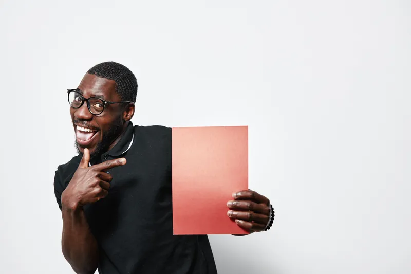 African American man with dark skin and glasses holding red paper pointing finger smiling on white background. Casual clothing, happy expression and studio portrait with copy space.