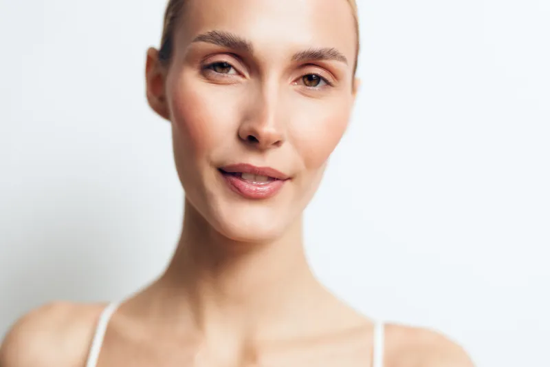 Close-up portrait of a young woman with smooth skin and a soft smile, facing the camera against a plain white background, conveying calm confidence and natural beauty