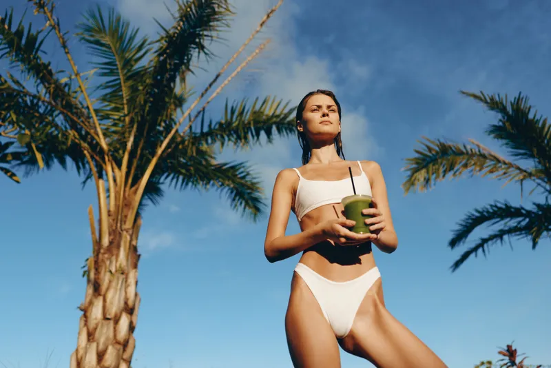 Active woman in a white bikini stands under palm trees, holding a coconut drink. Sunny outdoor scene emphasizes tropical lifestyle, summer wellness, and carefree leisure.
