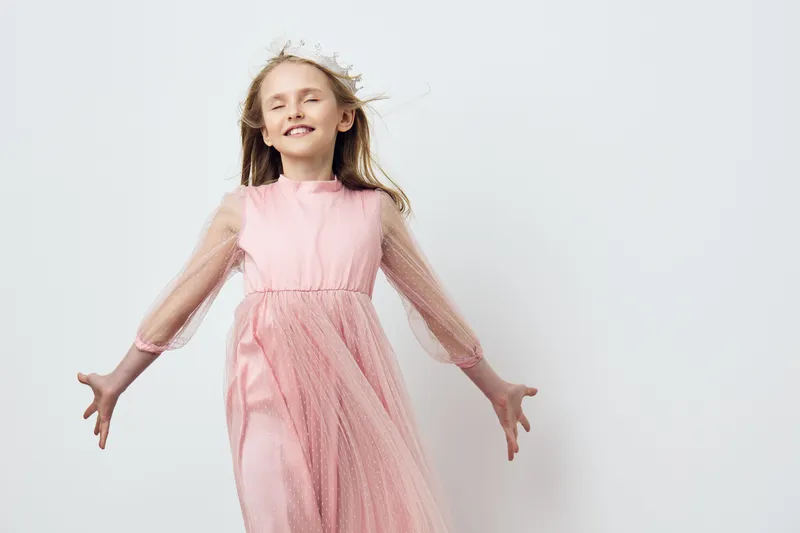 A joyful young girl wearing a pink dress stands against a neutral background, radiating happiness and innocence with outstretched arms