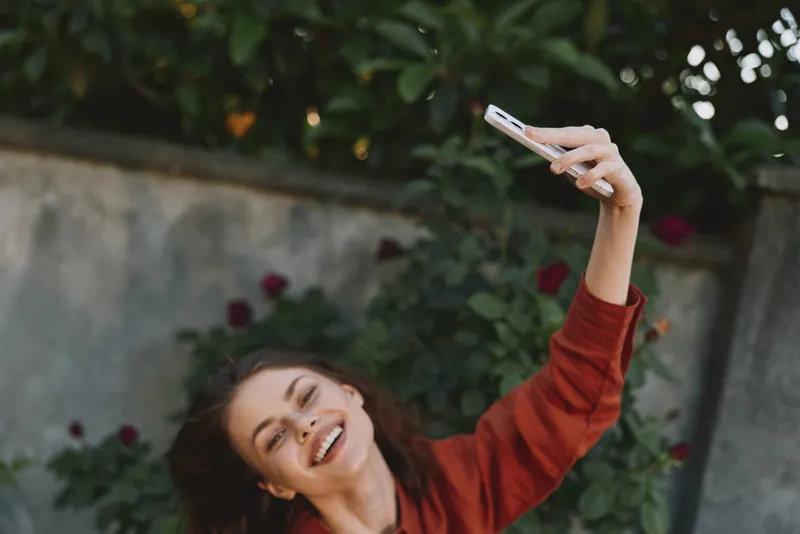 Cheerful woman outdoors, smiling while taking a selfie, wearing a red sweater, in a sunlit garden setting, casual lifestyle, candid moment, natural tones and relaxed posture