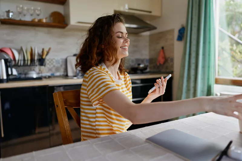 Woman enjoys a candid moment at home in a sunny kitchen, smiling while reaching across a table, casual and relaxed lifestyle scene with natural daylight.