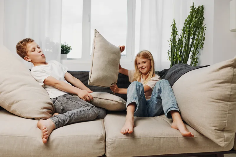 Two children playfully fight with pillows on a beige sofa in a bright living room with large windows and green plants. The moment captures fun, childhood, and sibling interaction.