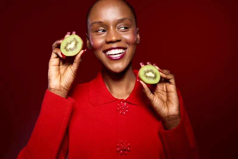 Smiling dark skinned African woman in red sweater holds two halves of fresh kiwi fruit near face, isolated on red background. Concept of healthy lifestyle, fruit, wellness, and natural beauty.