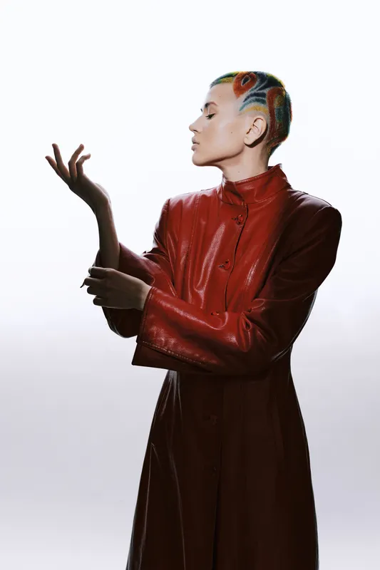 Woman with colorful patterned hairstyle wearing a shiny red leather coat, posing elegantly against a light background, showcasing modern fashion and bold style.