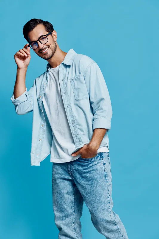 Stylish smiling young man in glasses wearing light denim shirt and white t-shirt poses confidently against solid blue background, demonstrating casual fashion concept.
