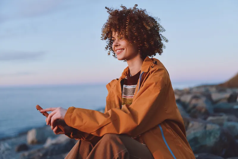 Woman lifestyle outdoors by sea, smiling, wearing orange jacket, curly hair, wind tousling, warm sunset light, relaxed body pose, coastal rocks background, calm natural colors, breeze