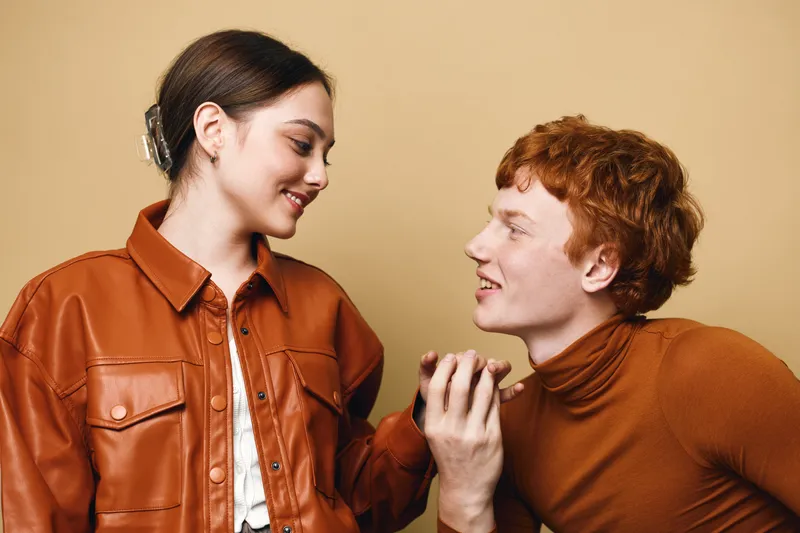Young couple in brown clothing looking at each other with smiles indoors on beige background. Attractive man and woman expressing affection and connection in casual style.