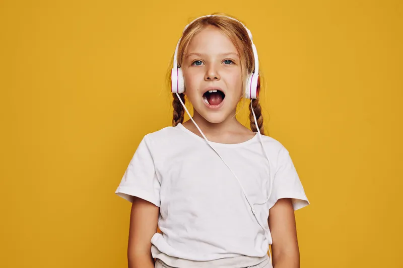 Happy girl with braids wearing headphones singing or speaking on yellow background. Casual white t-shirt and fun expression.