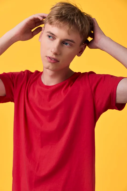 Young man in a red shirt, hands at his head, against a bright yellow backdrop, conveys a contemplative, casual mood and modern lifestyle energy in a studio scene