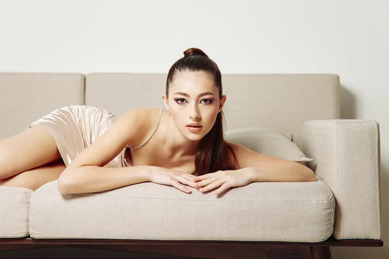 Young woman in creative fashion posing on beige couch with glossy dress and bold makeup, showcasing modern beauty style in indoor minimalistic setting.