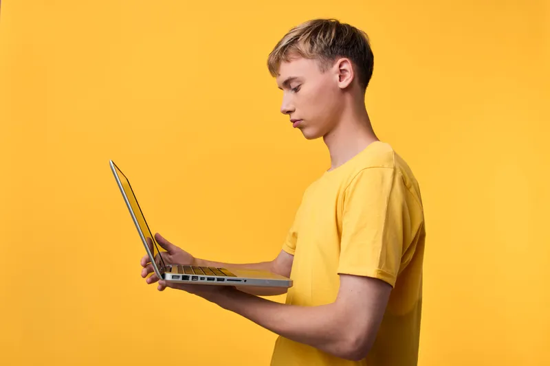 Young man in a yellow shirt studies on a laptop, standing against a vivid orange background, focusing on screen work, digital learning, and modern tech usage for online tasks