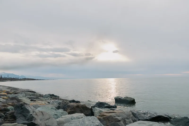 Rocky shore under cloudy sky with calm sea and soft sunlight reflecting on water surface. Peaceful coastal landscape with gentle waves and distant mountains in hazy background.