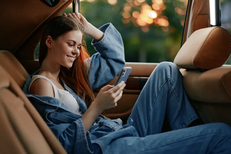 Smiling young woman in casual denim outfit using smartphone in a car during sunset, radiating happiness and relaxation.