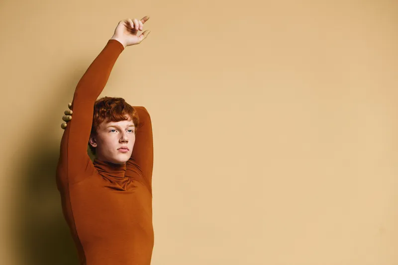 Young man with red hair and freckles stretching arms up wearing orange long sleeve sweater standing against plain beige background in studio. Relaxation, casual style and isolated portrait.