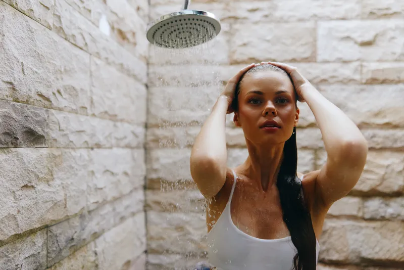 Woman taking outdoor shower with water droplets, wearing white tank top, natural stone wall background, wet hair falling, serene expression in warm daylight.