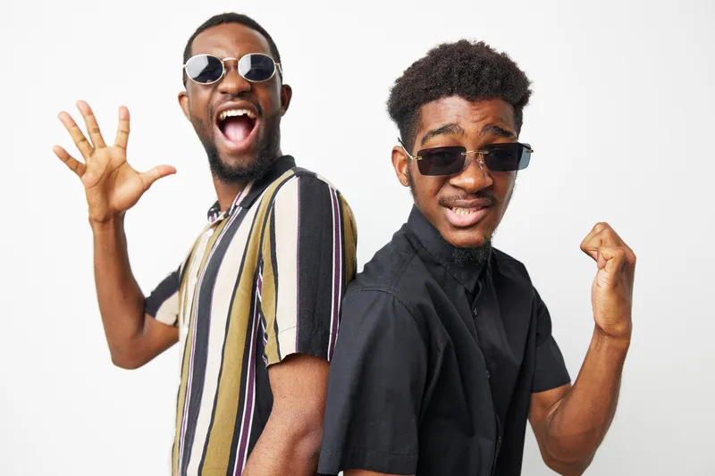 Two stylish Black men posing confidently with sunglasses, expressing joy and excitement. Their vibrant outfits add to the lively atmosphere of the image.