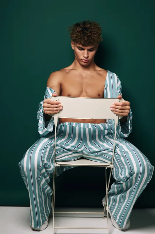 Young man with curly hair wearing striped pajamas sits on chair backwards against dark background, looking down with serious expression in studio portrait.