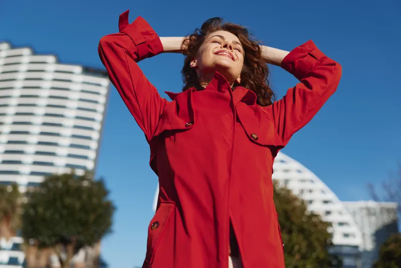 Cheerful woman outdoors in a red coat, smiling in sunlight, relaxed pose, gentle breeze, urban backdrop with modern buildings, vivid colors