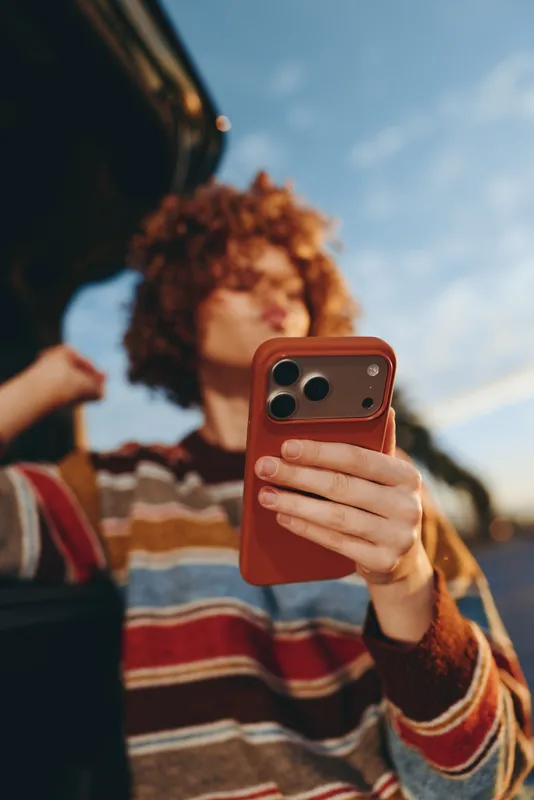 Woman with curly hair holding smartphone for a casual selfie outdoor. Warm evening light, soft blur background, striped sweater, lifestyle portrait, candid everyday mood, smile.