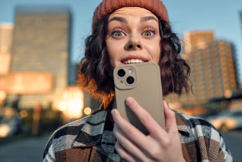 Woman with smartphone taking a selfie in urban city streets at golden hour, candid authenticity and emotion in a surprised smile wearing beanie, mindful living and emotional storytelling.