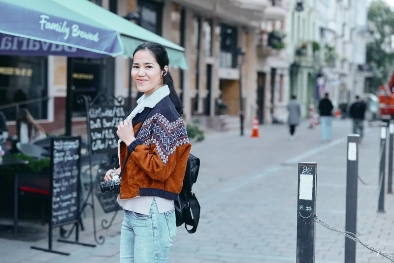 Street scene with a woman wearing a patterned jacket and light jeans, carrying a backpack, smiling softly as she strolls past cafe terraces on a sunny urban day.
