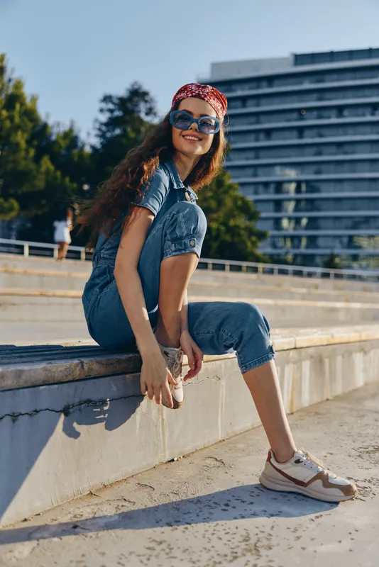 Lifestyle woman in modern boho-western denim street style wearing sunglasses and a red bandana, smiling outdoors in warm natural light with film color effect.