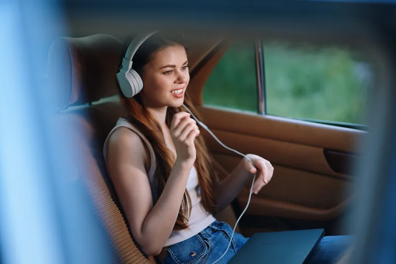 Young woman listening to music in a car, expressing happiness and relaxation, wearing casual clothes, with a vibrant green background and seasonal feel.