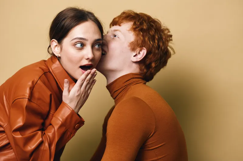 Whisper, secret, young man and woman with surprise and emotion in casual clothes on beige background. Communication, intimacy and reaction of friends sharing confidential message.