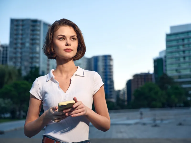 A cheerful young woman stands outdoors in an urban setting, gazing ahead while holding a smartphone, radiating calm confidence and a modern lifestyle.