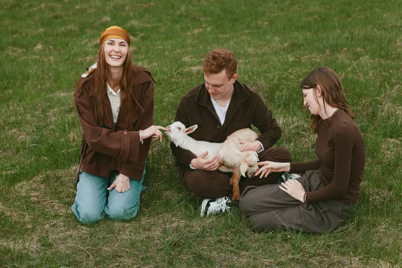 A warm outdoor scene features three friends kneeling on grass, gently petting a curious goat kid, capturing a lifestyle moment of friendship, care, and playful bond with animals in nature.