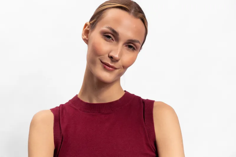 Young woman with light skin and brown hair wears sleeveless burgundy top, smiling softly on white background. Portrait of confident relaxed female model with natural makeup and casual style.